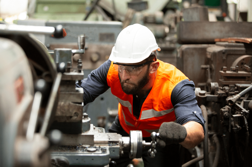 A man in an orange vest performs maintenance on a machine, focusing on safety equipment.