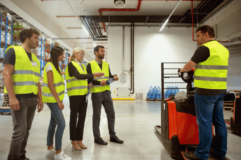 A group of people participating in safety training in a warehouse, surrounded by forklifts and safety equipment.